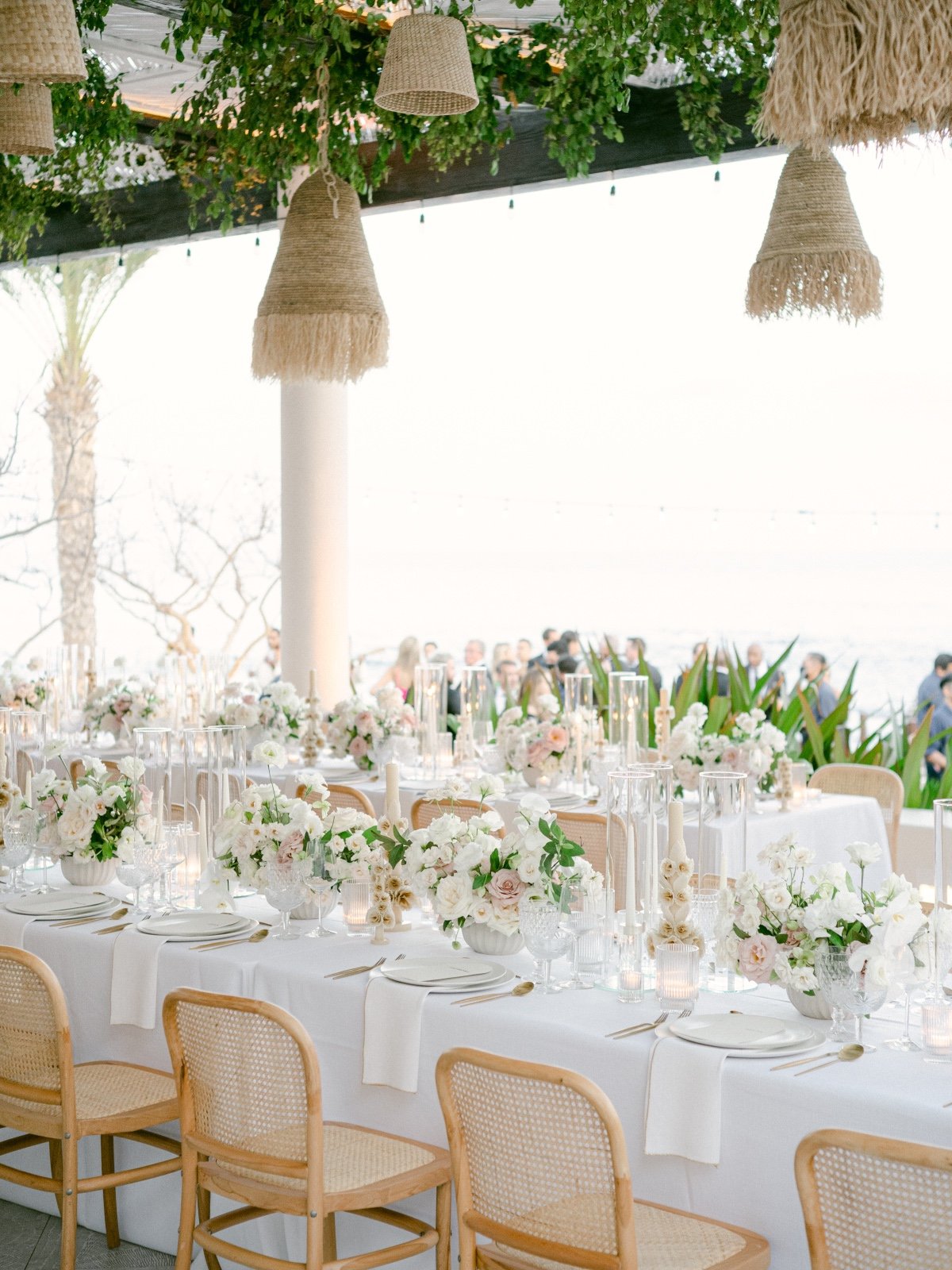 Reception table with white and pink floral arrangements, rattan chairs, and candles under glass