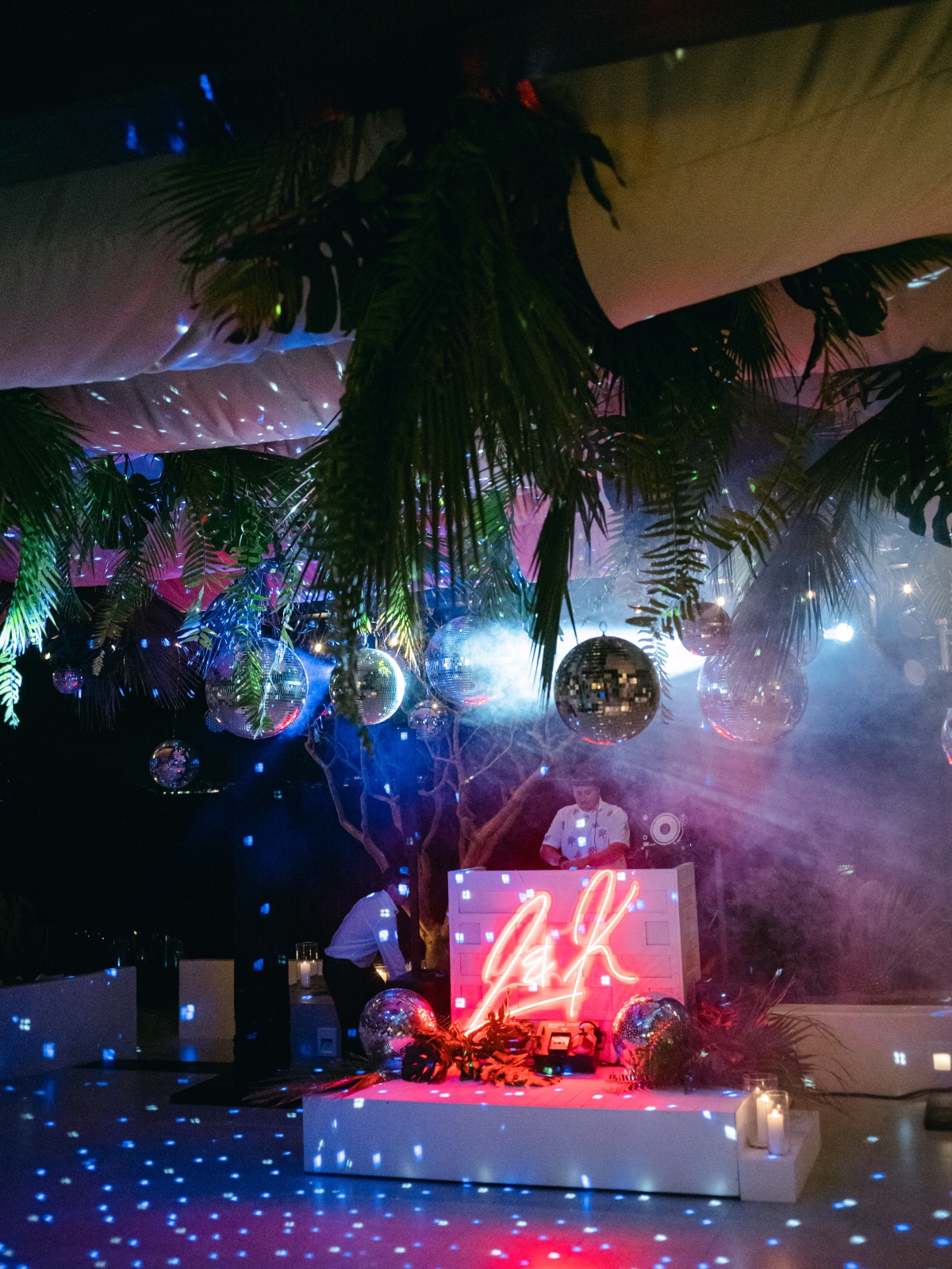 DJ behind booth with custom neon sign, tropical leaves, and disco balls