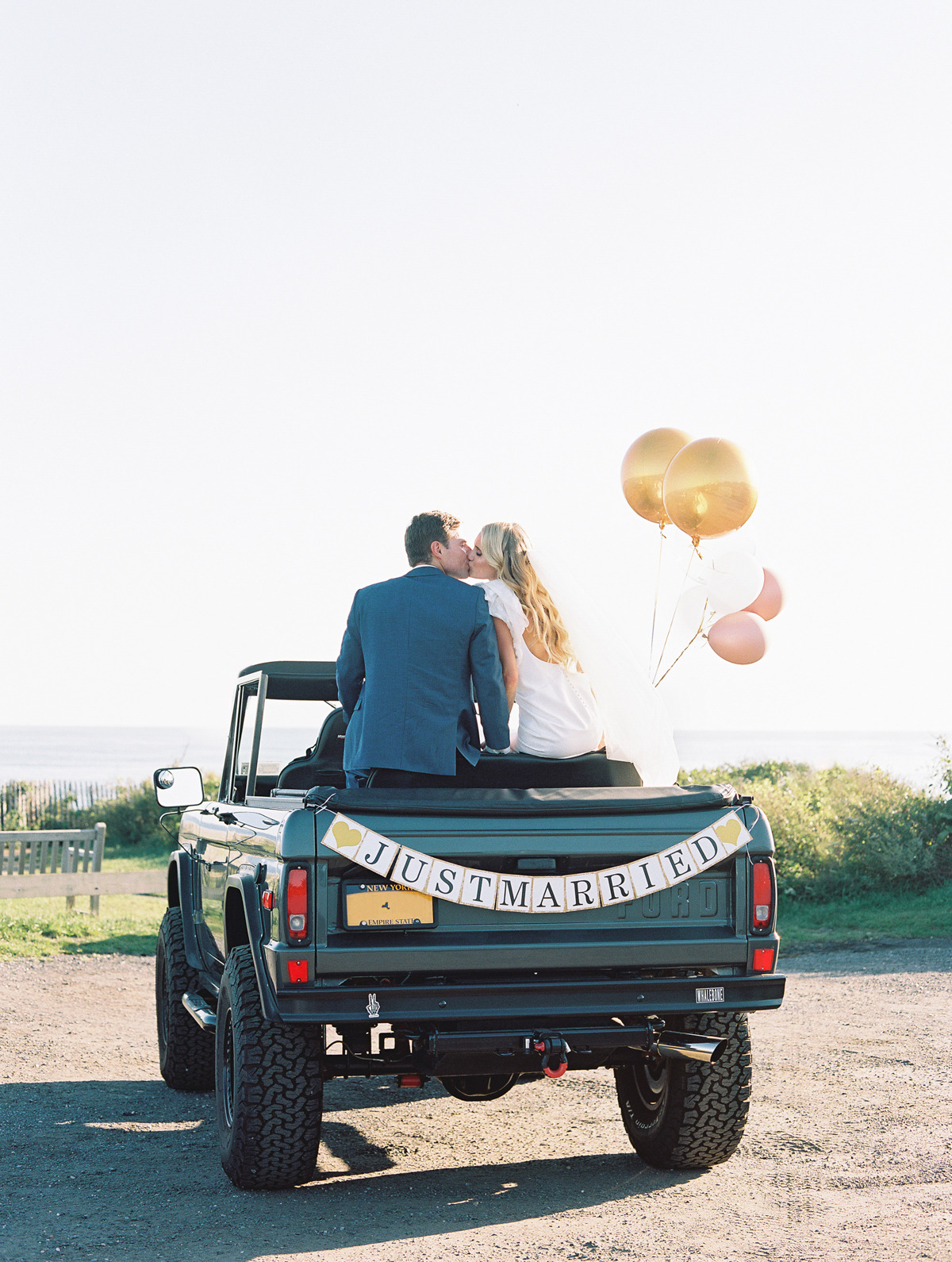 Bride and groom kissing on back of car with balloons and sign