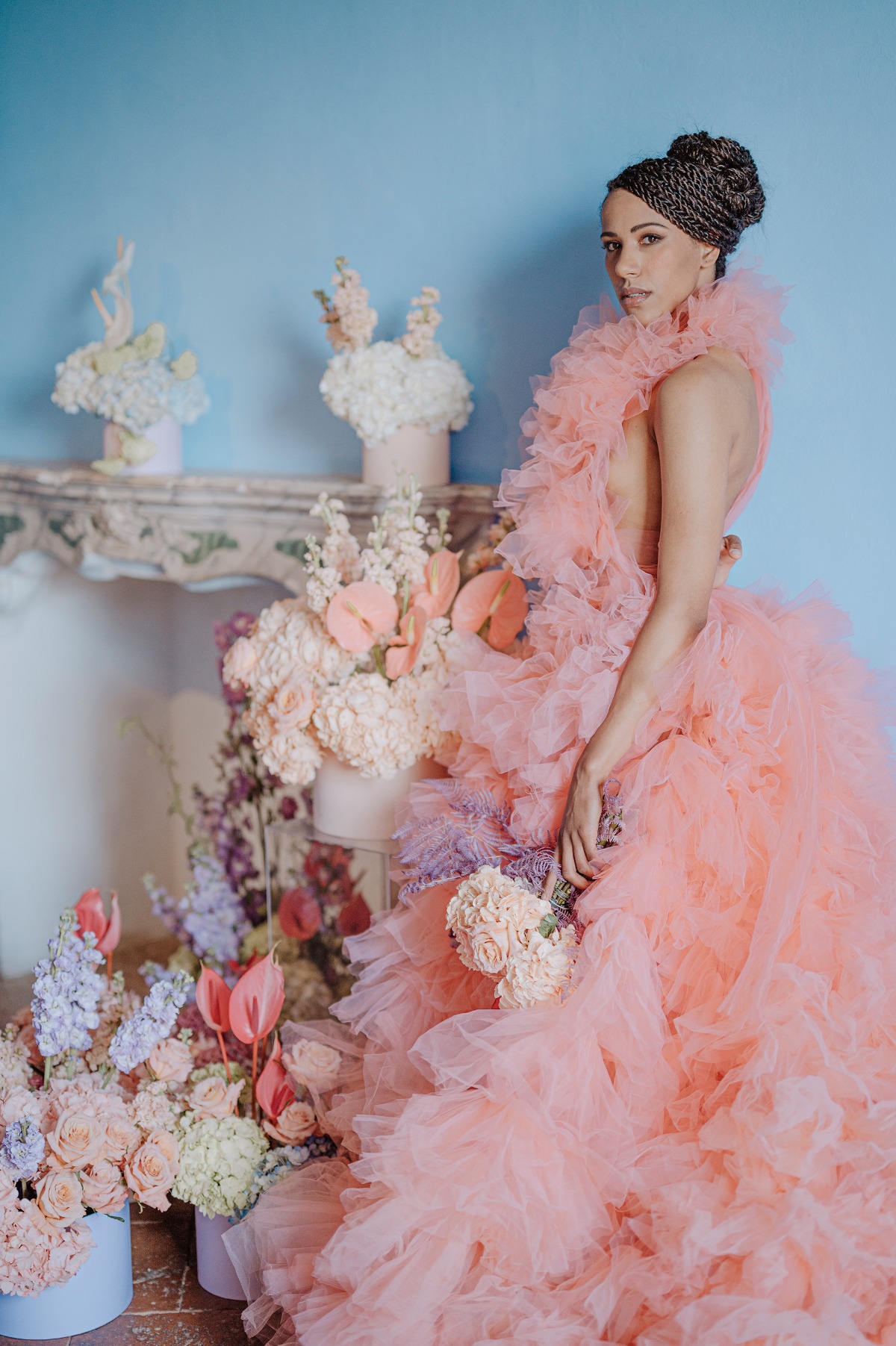 Bride in orange ruffled dress surrounded by florals in front of blue wall