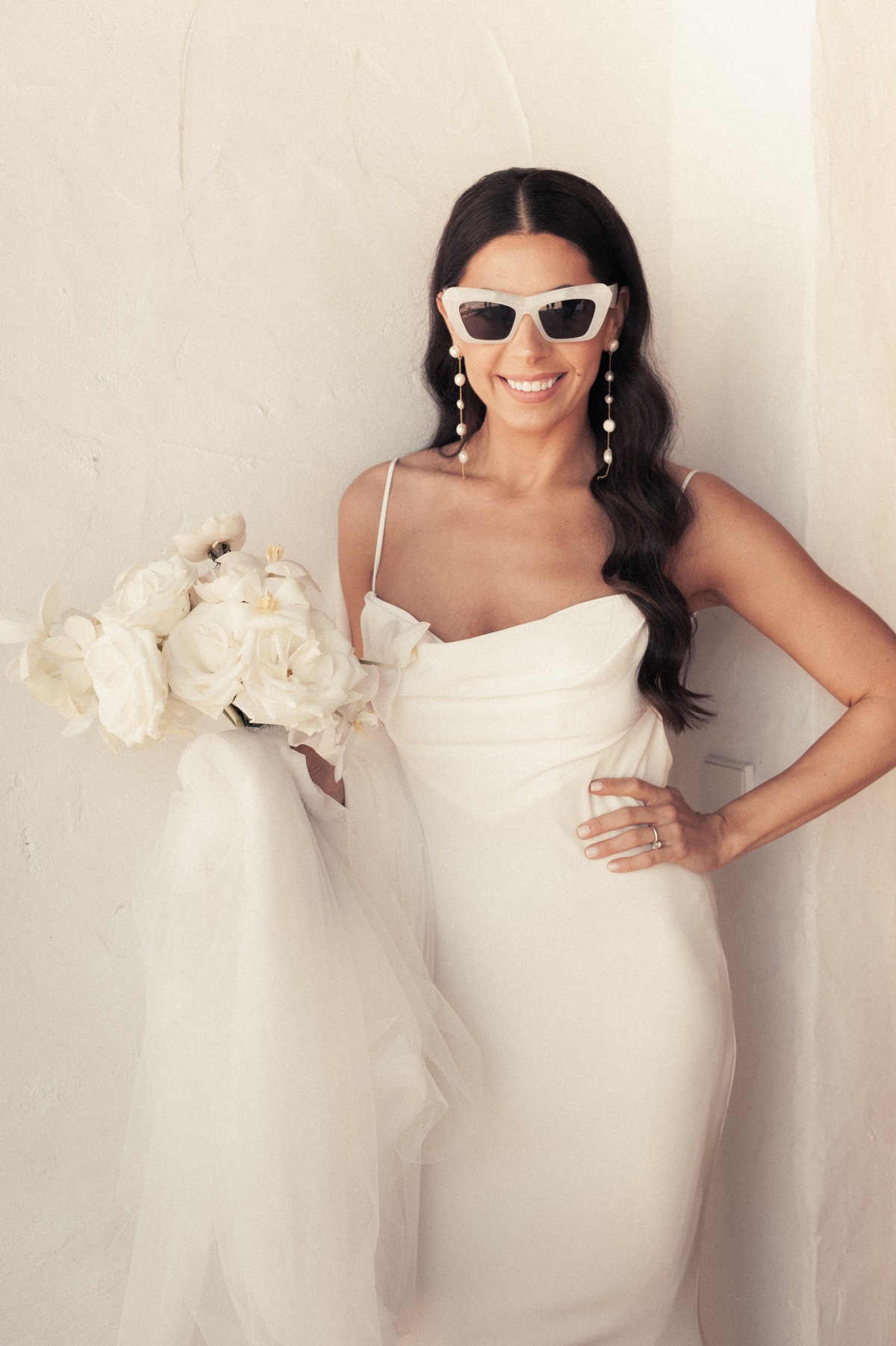 Bridal portrait holding bouquet and wearing sunglasses against white wall