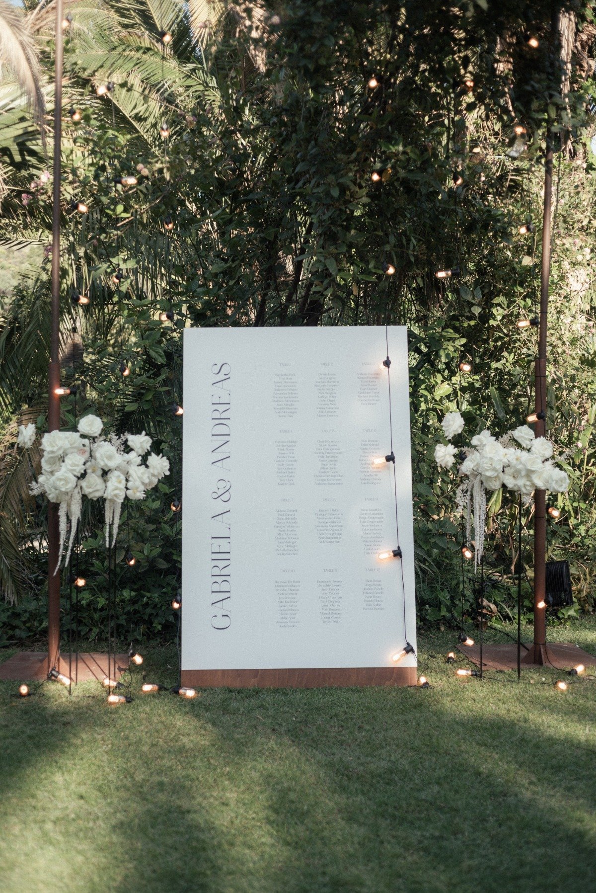 Reception seating chart behind string lights surrounded by white floral arrangements