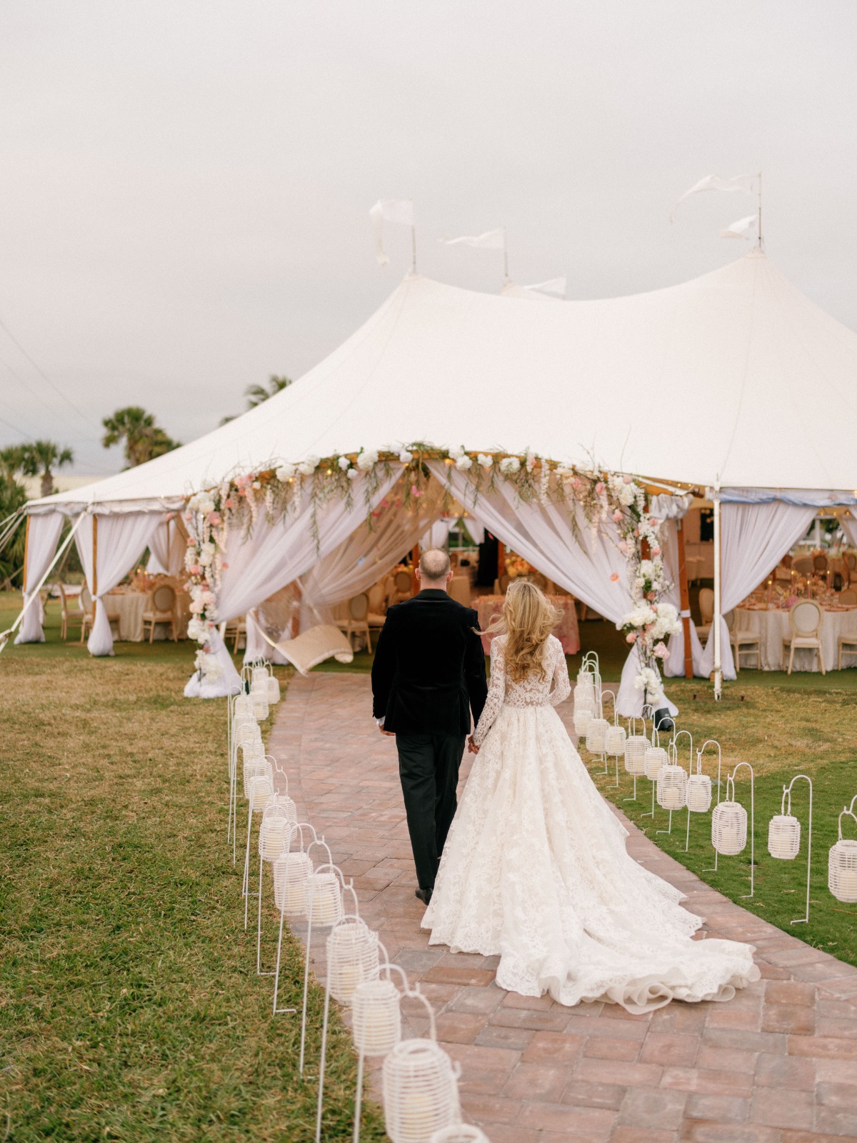 Bride and groom walking down pathway lined with lanterns into reception tent