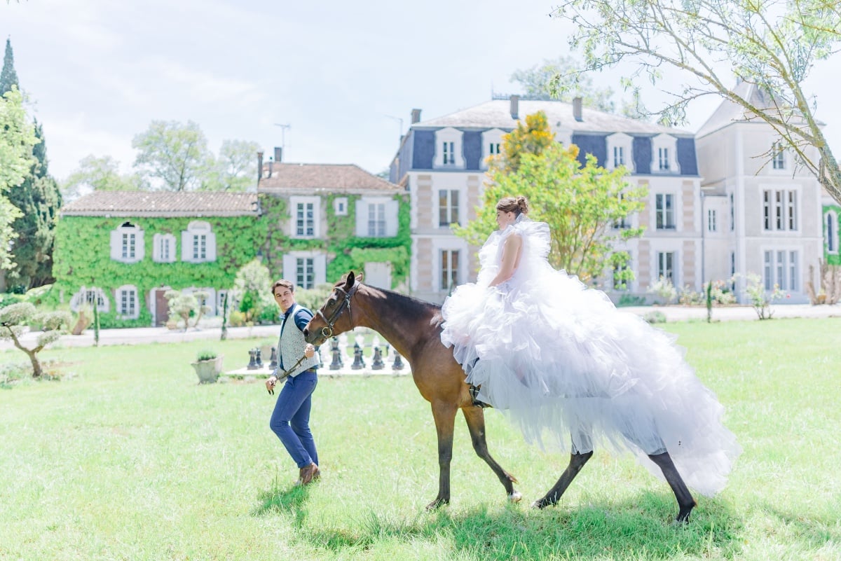 Bride riding horse in front of estate guided by Prince Charming