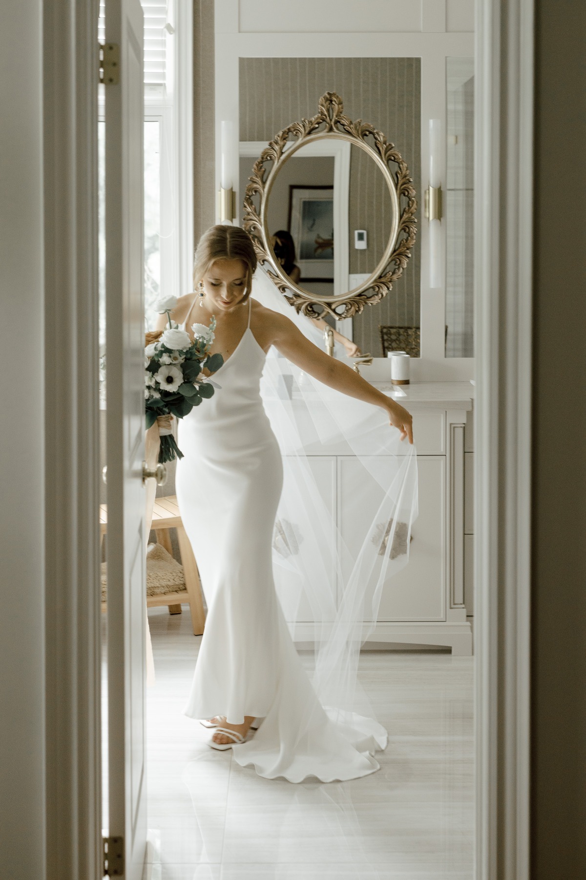 Bridal portrait in front of ornate bathroom mirror holding bouquet and veil