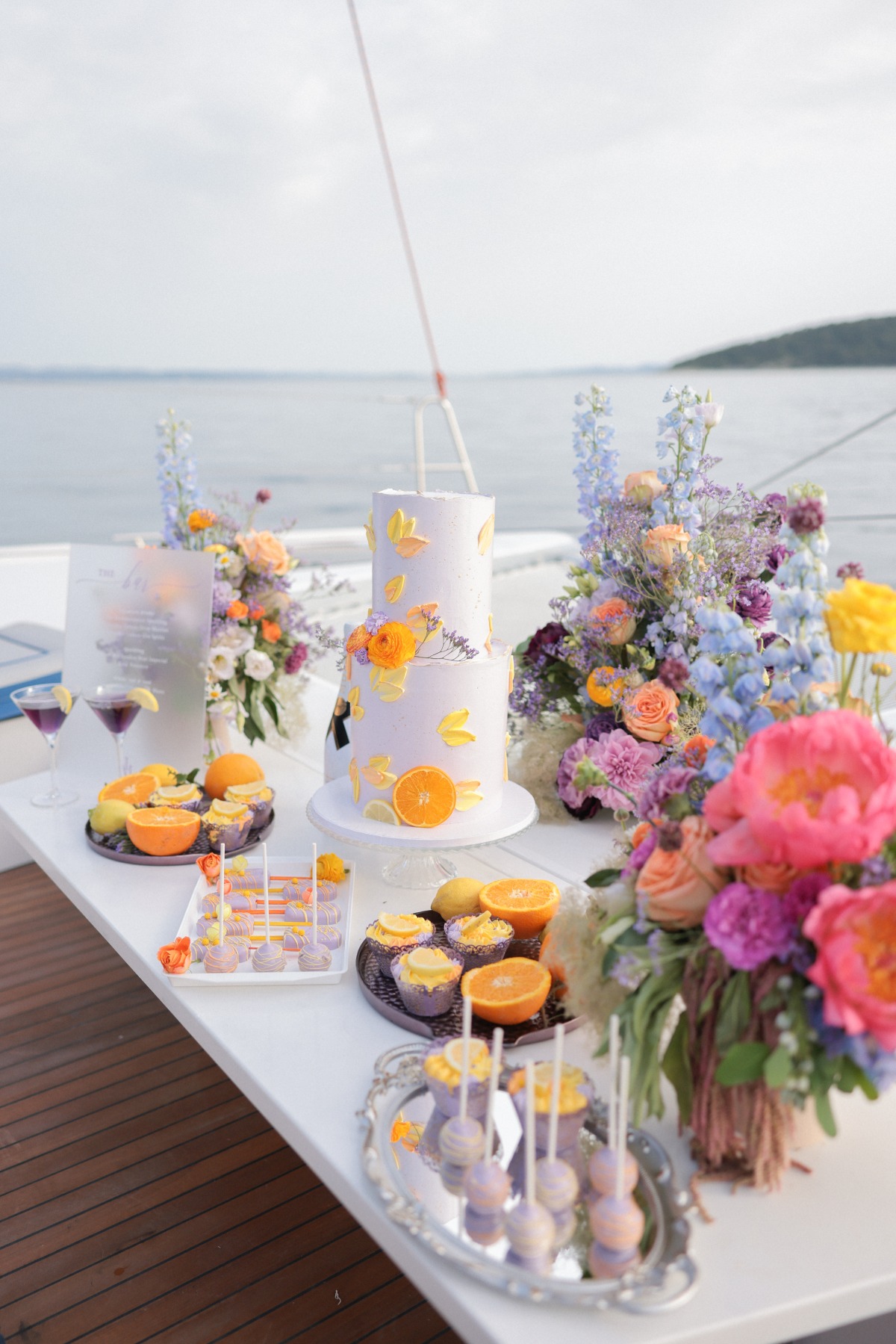 Dessert table on yacht with customized drinks, colorful arrangements, and floral cake