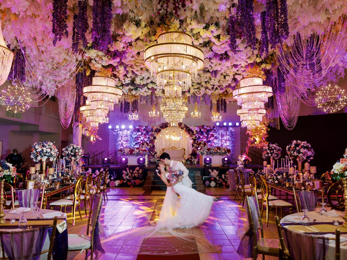 Groom dipping and kissing bride in ornate reception hall with chandeliers and flower ceiling piece