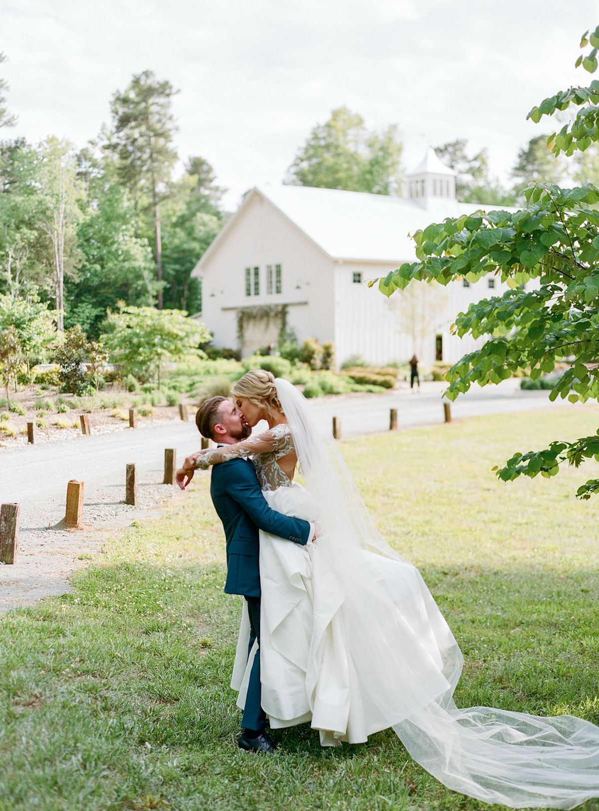 Groom holds bride in air while kissing her with barn in background