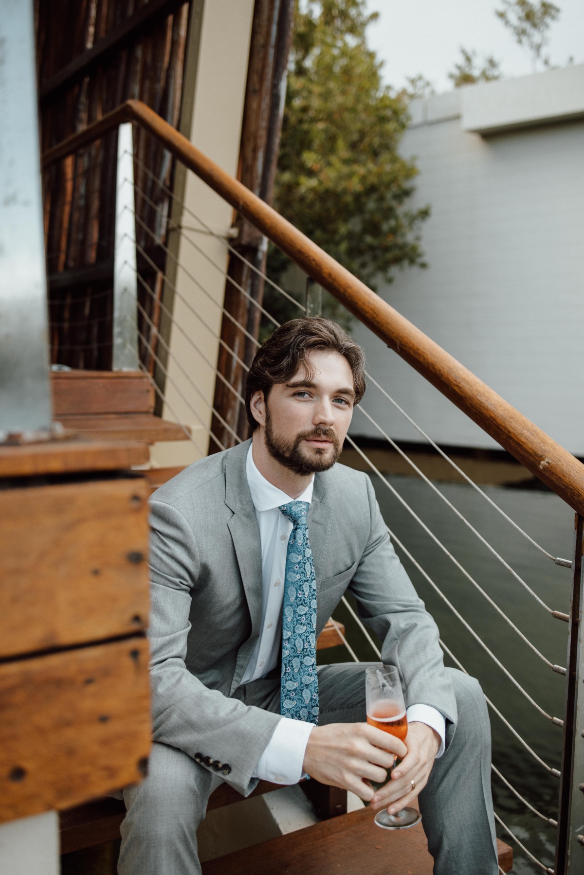 Groom in grey suit sitting on steps holding a glass of champagne