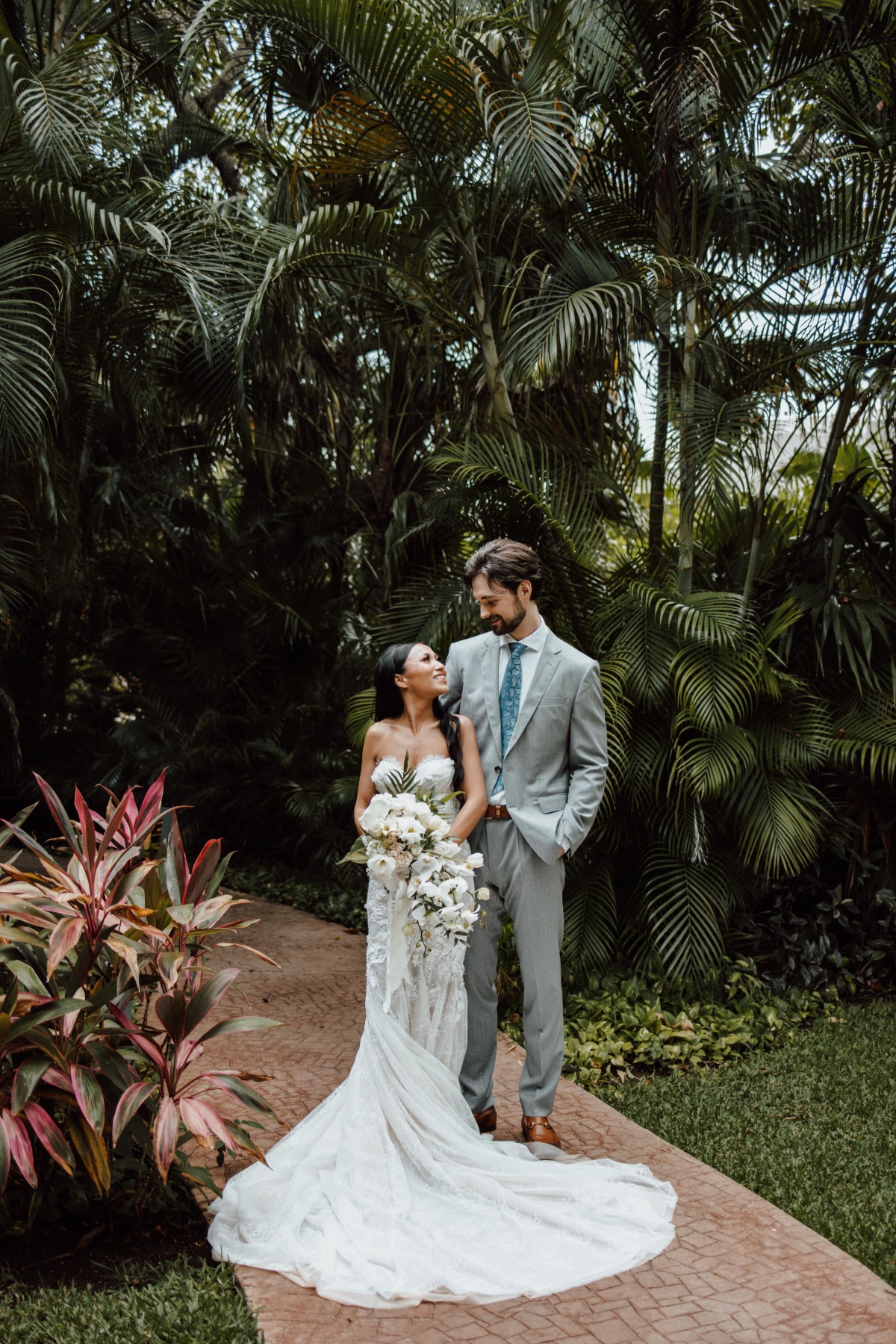 Bride and groom in front of tropical foliage holding white waterfall bouquet of white flowers