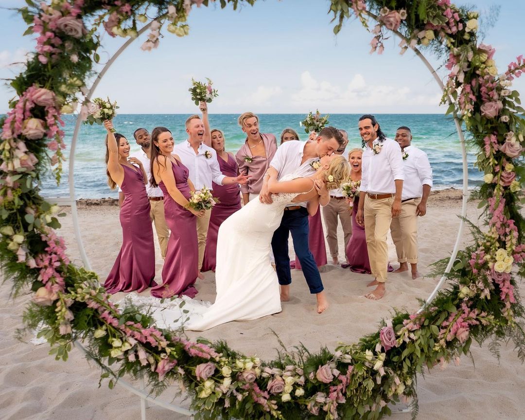 Newlyweds share a kiss on a beach wedding ceremony, surrounded by friends