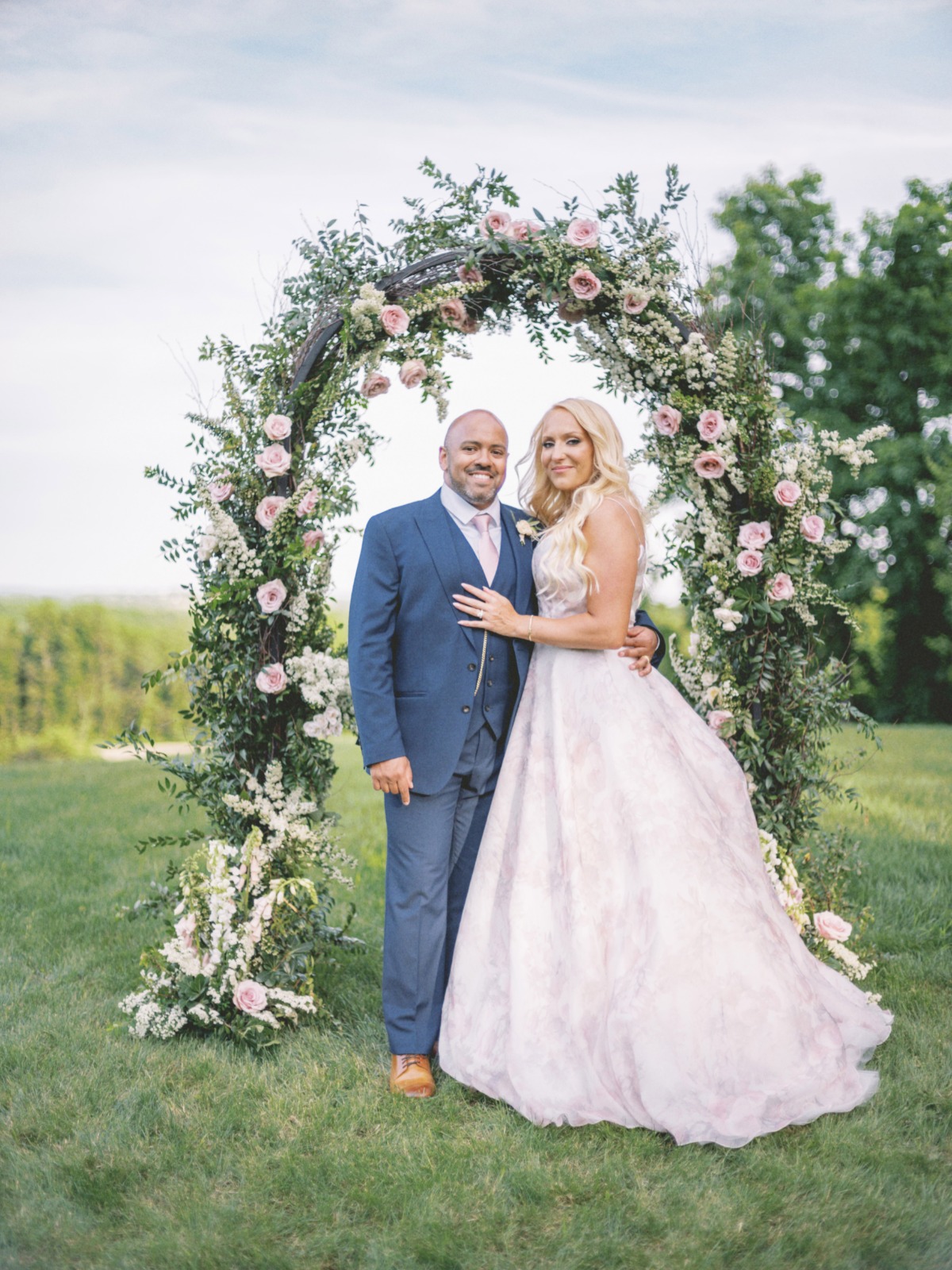 bride in blush floral dress with groom in blue suit in front of floral arch