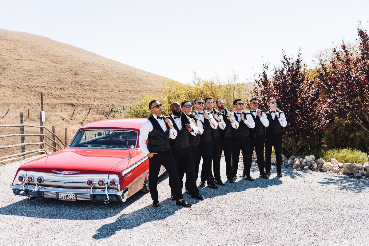 groomsmen in three piece tuxes standing in front of red low rider vintage car