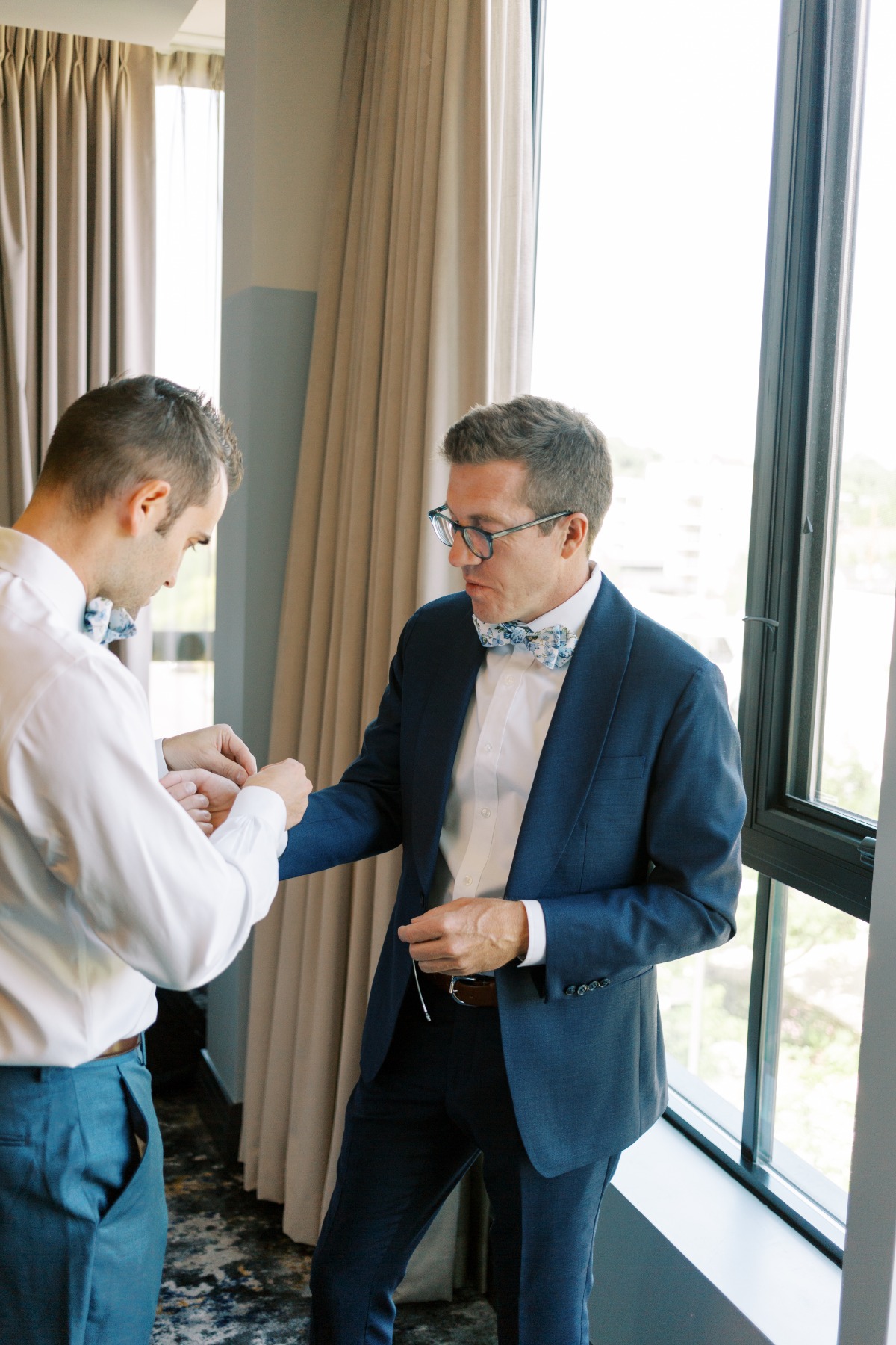 Groom helping other groom put on blue floral bow tie & cufflinks
