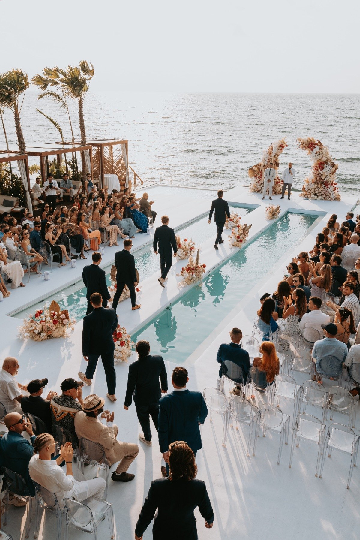 groomsmen walk down the aisle over pool at beach wedding